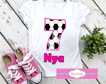 May include: White t-shirt with a pink soccer ball design in the shape of the number 7. The name "Nyaa" is written below the number in pink. The shirt is on a white wooden surface.