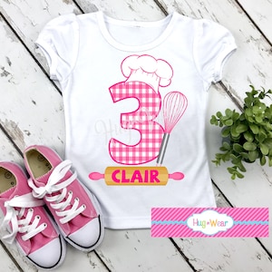 Op de afbeelding: Wit T-shirt met een roze en wit geruit nummer 3, een roze koksmuts, een roze garde en een deegroller met het woord "CLAIR" erop. Het shirt heeft een roze en blauw gestreept label met de tekst "Hug*Wear".