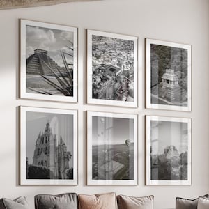 Könnte beinhalten: Sechs gerahmte Schwarz-Weiß-Fotografien von architektonischen Wahrzeichen hängen an einer beigefarbenen Wand. Die Fotografien zeigen antike Ruinen, Stadtlandschaften und Gebäude. Vor der Wand befindet sich ein braunes Ecksofa mit Kissen und ein runder Couchtisch.