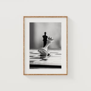 Puede incluir: Fotografía en blanco y negro enmarcada de una partida de ajedrez. El rey blanco ha caído sobre el tablero de ajedrez, mientras que el rey negro permanece de pie. La fotografía es en blanco y negro.