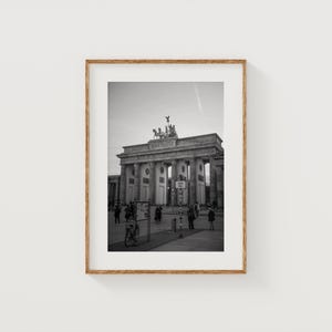 May include: Black and white framed photograph of the Brandenburg Gate in Berlin, Germany. The neoclassical monument is captured from a ground-level perspective, with people and a bicycle in the foreground. The sky is overcast.