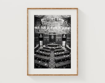 Könnte beinhalten: Schwarz-Weiß-Fotografie eines großen Raumes mit Reihen von Schreibtischen und Stühlen, wahrscheinlich ein Parlamentssaal. Ein großer Kronleuchter hängt von der Decke. Das Bild ist in einem Holzrahmen gerahmt.