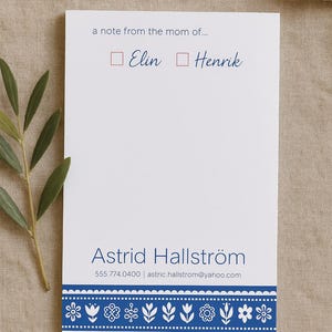 May include: A white notecard with a blue border and a floral design. The text "a note from the mom of..." is printed at the top. There are two checkboxes with the names "Elin" and "Henrik" written next to them. The bottom of the card has the name "Astrid Hallström" and contact information.