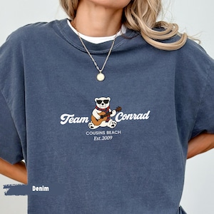 May include: Denim blue t-shirt with the text "Team Conrad" and a graphic of a bear wearing sunglasses and playing a guitar. The text "Cousins Beach Est. 2009" is below the bear graphic.