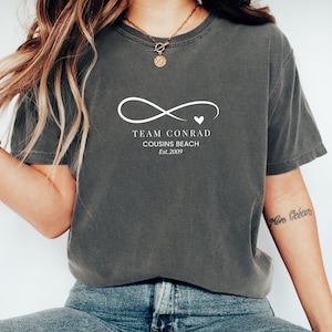 May include: Dark gray t-shirt with the text "TEAM CONRAD Cousins Beach Est.2009" in white. The design includes an infinity symbol with a heart. The shirt is a casual wear item.