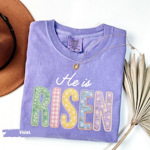 Könnte beinhalten: Ein violettes Comfort Colors T-Shirt mit den Worten "He is RISEN" in bunten Buchstaben im Patchwork-Stil. Eine goldene Halskette mit Anhänger liegt auf dem Shirt. Ein brauner Hut und ein getrocknetes Palmblatt sind im Hintergrund.