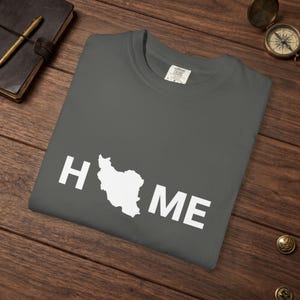May include: A charcoal gray crew neck t-shirt featuring the white text "HOME" printed on the front, where the letter "O" is replaced by a white silhouette map of Iran. The shirt has a visible interior neck label with the brand name Comfort Colors.