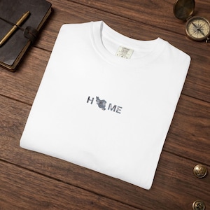 May include: White t-shirt with the word "HOME" printed in a dark gray, stylized font. The "O" in "HOME" is replaced with a dark gray graphic of a country's map. The shirt is folded on a wooden surface.