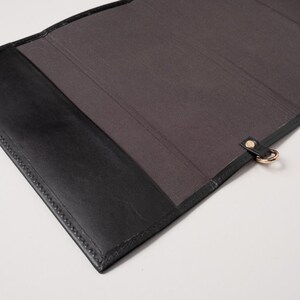 May include: Black leather folio with a dark grey interior. The folio has a stitched border and a gold-coloured metal ring attached with a leather strap. The folio is open, revealing the interior.