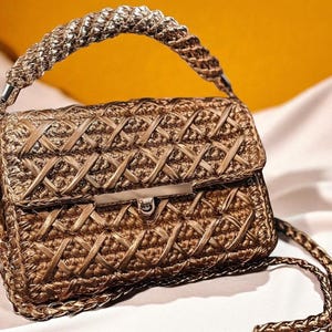 May include: A gold-toned handbag with a woven design. The bag features a braided handle and a chain strap. The flap closure has a silver-colored clasp. The bag's texture and color give it a luxurious appearance.