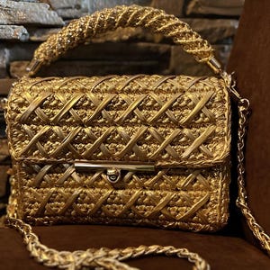 May include: A gold-colored handbag with a woven design. The bag features a braided handle and a chain strap. The clasp is a gold-toned metal. The bag is sitting on a brown surface.