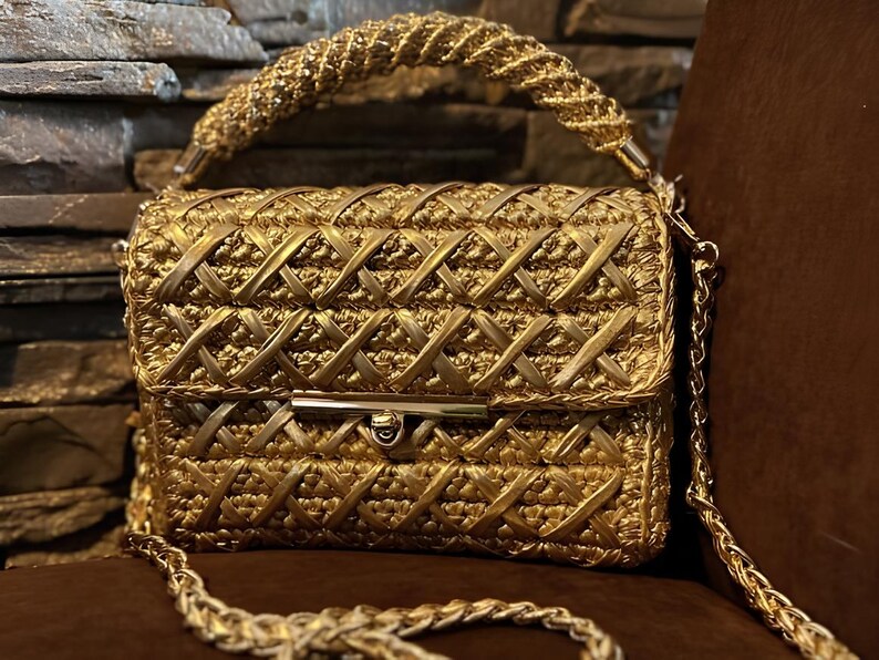 May include: A gold-colored handbag with a woven design. The bag features a braided handle and a chain strap. The clasp is a gold-toned metal. The bag is sitting on a brown surface.