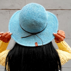May include: A light blue woven sun hat with a brown band and a small brown tag. The hat is held up by a person wearing a yellow sweater. The background is a gray wall.
