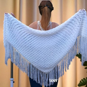 May include: A white, triangular shawl with a textured knit pattern and long fringe. The shawl is held up, displaying its full shape and design. The fringe hangs down along the edges, adding a decorative element to the garment.