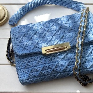 May include: A light blue, handmade crochet handbag with a diamond pattern. The bag features a gold-colored clasp and chain strap. The handle is also crocheted. The bag is on a white wooden surface.