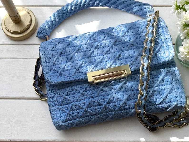 May include: A light blue, handmade crochet handbag with a diamond pattern. The bag features a gold-colored clasp and chain strap. The handle is also crocheted. The bag is on a white wooden surface.