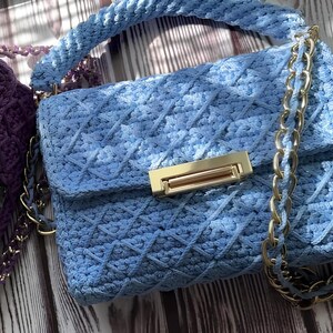 May include: A light blue, handmade crochet handbag with a gold-tone clasp and chain strap. The bag features a diamond pattern and a matching braided handle. A purple crochet bag is partially visible in the background.