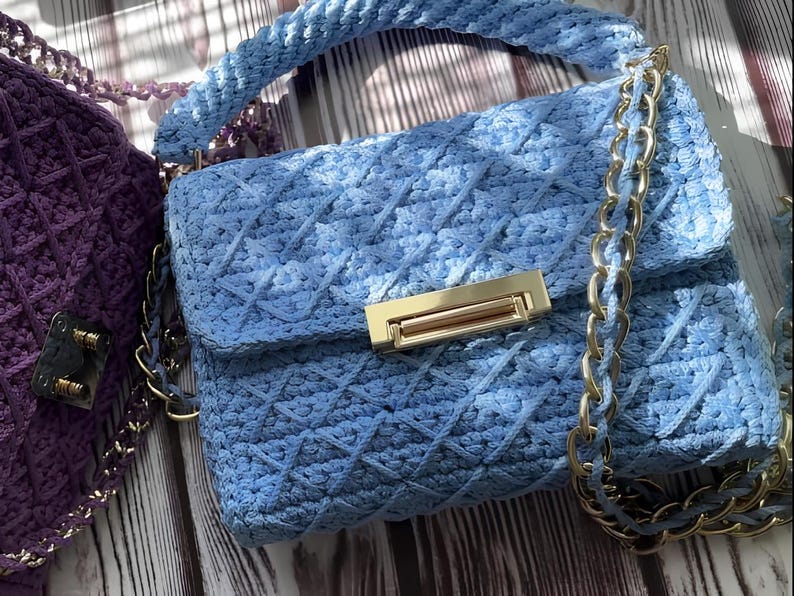 May include: A light blue, handmade crochet handbag with a gold-tone clasp and chain strap. The bag features a diamond pattern and a matching braided handle. A purple crochet bag is partially visible in the background.
