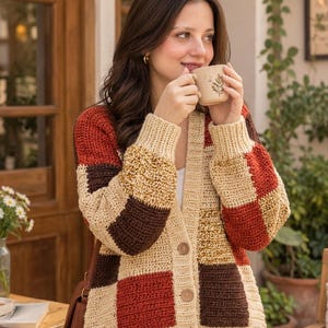 May include: A long-sleeved, button-down cardigan featuring a patchwork design of square blocks in cream, dark brown, and rust orange. The garment is constructed with a textured crochet stitch, ribbed cuffs, and a ribbed hem, finished with three buttons.