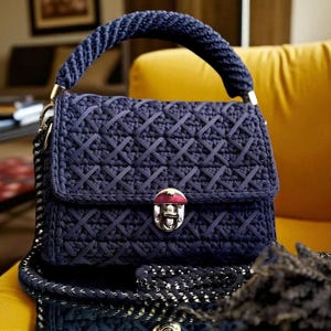 May include: A navy blue handbag with a woven pattern, a braided handle, and a silver clasp. The bag has a flap closure and a chain strap. The bag is sitting on a yellow surface.