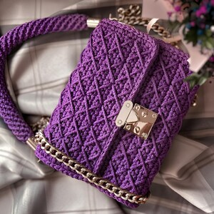 May include: A handmade, purple crochet handbag with a geometric pattern. The bag features a braided handle, a gold-colored chain strap, and a gold-tone clasp. The bag is approximately 10 inches wide and 7 inches tall.