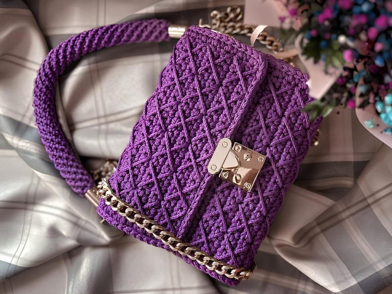 May include: A handmade, purple crochet handbag with a geometric pattern. The bag features a braided handle, a gold-colored chain strap, and a gold-tone clasp. The bag is approximately 10 inches wide and 7 inches tall.
