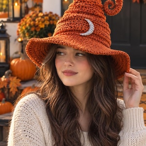 Puede incluir: Un sombrero de ganchillo color naranja quemado con ala ancha y una punta alta y curvada. El sombrero presenta un adorno de media luna plateada fijado en el centro de la parte delantera. La textura se compone de filas de puntos densos.