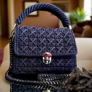 May include: A navy blue handbag with a woven pattern and a matching braided handle. The bag features a silver clasp and chain strap. The bag is sitting on a white surface.