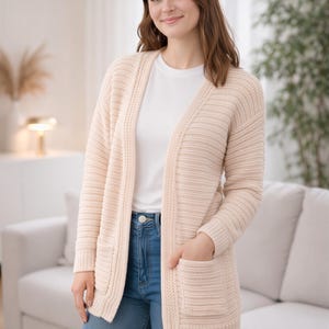 May include: A light beige open-front cardigan featuring a ribbed knit texture, long sleeves, and two front patch pockets. The garment is worn over a plain white crew-neck t-shirt and blue denim jeans, showcasing a simple and functional design.