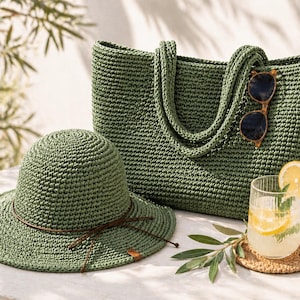 May include: A green crochet tote bag, sun hat, and a glass of lemonade with sunglasses. The tote bag has two handles and the hat has a brown band. The glass of lemonade has a lemon slice and mint leaves. The sunglasses are brown.