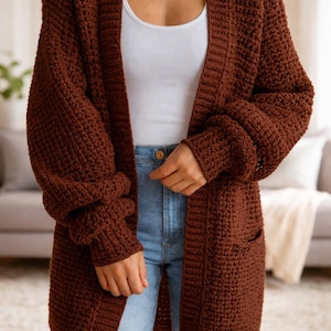 May include: A long, open-front, brown crochet cardigan with large pockets. The sleeves are long and cuffed. The cardigan is worn over a white vest top and blue jeans. The overall style is casual and comfortable.