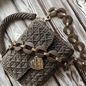 May include: A brown, woven handbag with a heart-shaped clasp. The bag features a chain strap with circular links and a braided handle. The bag is approximately 7 inches wide and 6 inches tall.