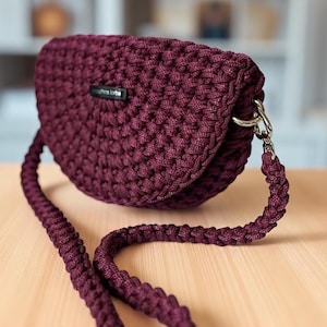 Puede incluir: Bolso bandolera de ganchillo burdeos, de forma redondeada, con una larga correa a juego. El bolso tiene un cierre plateado y una pequeña etiqueta rectangular. El bolso está hecho de un hilo grueso y texturizado.