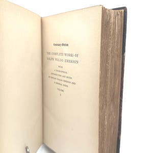Puede incluir: Un libro abierto con p&aacute;ginas blanquecinas envejecidas. El texto dice "Centenary Edition THE COMPLETE WORKS OF RALPH WALDO EMERSON" con texto adicional debajo. El lomo del libro es oscuro y los bordes de las p&aacute;ginas son visibles.