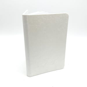 May include: A small, off-white journal with a textured cover stands open, revealing the white pages within. The book is upright, showcasing its spine and the edge of the pages. The cover has a subtle, leather-like grain.
