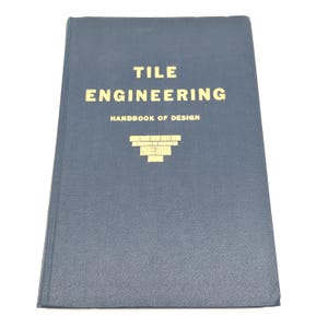 May include: A navy blue book titled "TILE ENGINEERING HANDBOOK OF DESIGN" with gold lettering and a gold graphic of a brick pattern. The book is closed and appears to be in good condition.