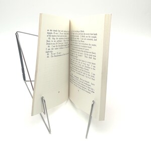 May include: A silver-colored metal book stand holding an open book. The book is white with black text. The stand has a simple, minimalist design, with two angled supports and a base.