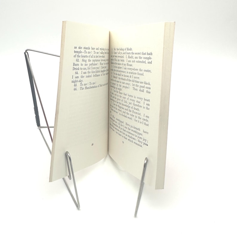 May include: A silver-colored metal book stand holding an open book. The book is white with black text. The stand has a simple, minimalist design, with two angled supports and a base.