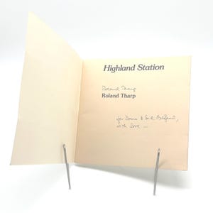 Può includere: Una carta aperta, color crema, con il testo "Highland Station" in grigio. Sotto il titolo ci sono i nomi "Roland Tharp" e "Roland Tharp". Il testo scritto a mano recita "for Donna & Sid Gelfand, with love -."