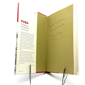 May include: A hardback book titled Cuba: The Measure of a Revolution by Lowry Nelson, featuring a white dust jacket with red text and a tan inner page with a handwritten black ink inscription signed by the author and dated 4-5-77.