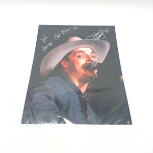 May include: A photograph of a man wearing a cowboy hat and denim shirt, singing into a microphone. The image has handwritten text that says "Val, Love ya. God Bless you!" and a signature with the year "97".