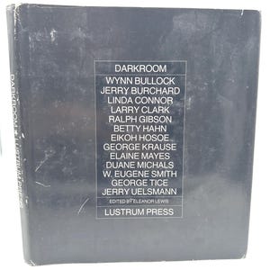 Puede incluir: Un libro gris oscuro titulado "DARKROOM" con una lista de nombres y "LUSTRUM PRESS" impreso en la portada. El lomo del libro también muestra el título y la editorial. El libro es probablemente un libro de fotografía.