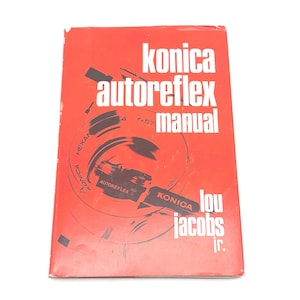 May include: A red Konica Autoreflex manual book with white text. The book cover features a camera lens diagram and the text "konica autoreflex manual" in large white letters. The author's name, Lou Jacobs Jr., is also visible.