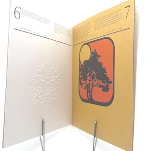 May include: Open book with pages displaying a snowflake design and a tree graphic. The left page features a white embossed snowflake. The right page has a tree silhouette within an orange and yellow rectangle. The book is held open by metal stands.
