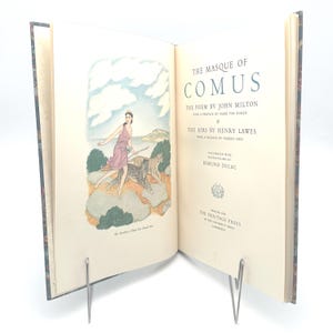 May include: Open book titled "The Masque of Comus" by John Milton, with illustrations by Edmund Dulac. The left page features a color illustration of a woman in a pink dress with a leopard. The book is held open by a metal book holder.