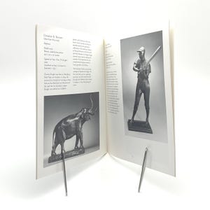 May include: An open book with two black and white images. One image is a bronze sculpture of an elephant, and the other is a baseball player holding a bat. The book is held open by two silver metal book holders.