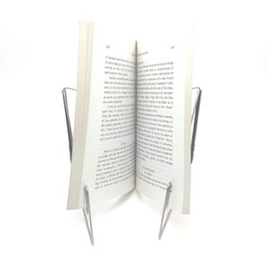 K&ouml;nnte beinhalten: Ein Buch, das von einem silbernen Metall-Buchst&auml;nder offen gehalten wird. Das Buch hat wei&szlig;e Seiten mit schwarzem Text. Der St&auml;nder hat ein schlichtes, minimalistisches Design.