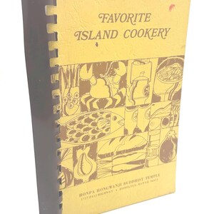 May include: A yellow spiral-bound cookbook titled Favorite Island Cookery. The cover features brown line art illustrations of various food items including a crab, fish, and vegetables. Text at the bottom reads Honpa Hongwanji Buddhist Temple.