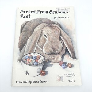 May include: A vintage book cover titled "Scenes From Seasons Past" by Claudia Nice, featuring a detailed illustration of a brown rabbit with a bowl of colorful marbles. The cover also includes the text "Presented By Sue Scheewe" and "vol. 7".