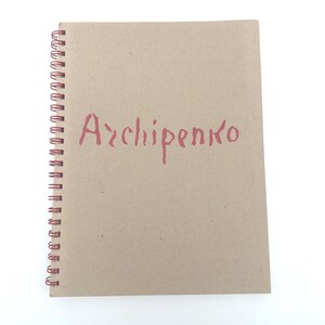 May include: A spiral-bound notebook with a kraft paper cover. The word "Archipenko" is printed in a reddish-pink color across the front. The spiral binding is also reddish-pink.